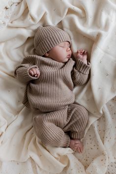 a newborn baby wrapped in a brown sweater and hat sleeping on a white bed sheet