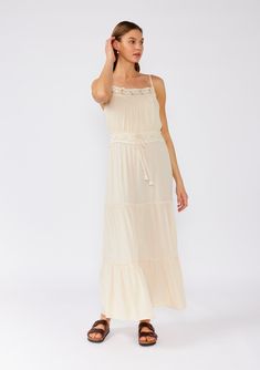 [Color: Natural] A front facing image of a brunette model wearing an off white sleeveless maxi dress. With adjustable spaghetti straps Bohemian Spaghetti Strap Dress With Crochet Trim, Summer Maxi Dress With Lace Trim And Tiered Skirt, Bohemian Dress With Crochet Trim And Spaghetti Straps, Summer Tiered Maxi Dress With Lace Trim, Bohemian Tiered Dress With Adjustable Straps, Bohemian Dress With Adjustable Straps And Tiered Design, Vacation Dress With Lace Trim And Tiered Skirt, Beach Maxi Dress With Crochet Trim, Vacation Maxi Dress With Lace Trim