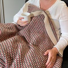 a woman sitting in a chair holding a coffee cup and blanket on her lap, smiling at the camera