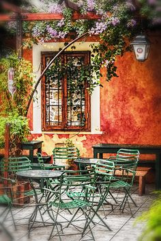 an outdoor dining area with green chairs and tables in front of a red wall that has flowers growing on it