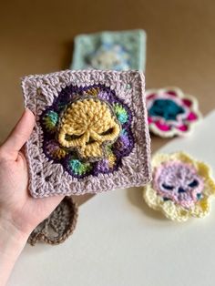 a hand holding up a crocheted square with a skull on it and three smaller squares in the background