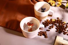 two coffee mugs with small figurines in them on a table surrounded by coffee beans