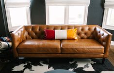 a brown leather couch sitting in front of two windows with pillows on top of it