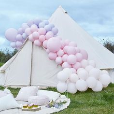 a teepee tent with pink and white balloons on the grass next to pillows, cake and cupcakes