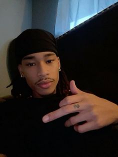 a young man with dreadlocks sitting on a couch pointing at the camera while wearing a black shirt