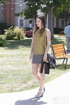 a woman standing on the sidewalk in front of a park bench and holding a purse