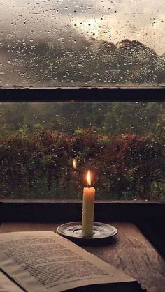 a candle is lit on a plate next to an open book and window with raindrops