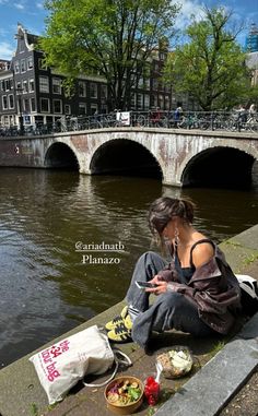 a woman sitting on the edge of a river looking at her cell phone and eating