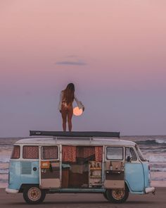 a woman standing on top of an old vw bus at the beach in front of the ocean