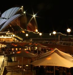 an outdoor restaurant with tables and umbrellas lit up at night in front of the sydney opera house