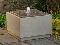 a water fountain in the middle of a stone walkway next to some plants and bushes