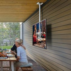 two people sitting at a picnic table with food on the outside patio, and one person standing up to eat