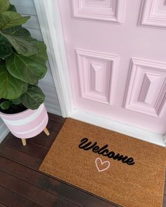 a welcome mat with a heart on it next to a potted plant in front of a pink door