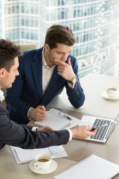 two men sitting at a table in front of a laptop computer looking at each other