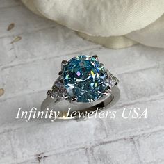 "The ring pictured is a simulated aquamarine with simulated diamonds #7034 -Approximate total carat weight: 5.00ctw diamond equivalent -Center Stone Size: 11x9mm - approx. 4.20ct diamond equivalent -Center Stone Shape: oval cut -Side Stones Size: 2 Trillions 5x5mm - approximately 0.80ctw diamond equivalent -Gem Type: simulated diamond -Stone Clarity: VVS1 -Stone Color: light blue center stone -Moh's Scale: 8.5 hardness -Metal Type and Purity: 14k white gold -Setting: 4 prong tulip head -Stock Ri Light Blue Oval Ring With Prong Setting, Light Blue Diamond Ring For Anniversaries, Oval Aquamarine Diamond Ring With Accents, Oval Aquamarine Diamond Ring With Diamond Accents, Gia Certified Oval Topaz Ring For Gift, Gia Certified Oval Topaz Ring As Gift, Anniversary Light Blue Aquamarine Diamond Ring, Oval Light Blue Promise Ring, Light Blue Oval Promise Ring