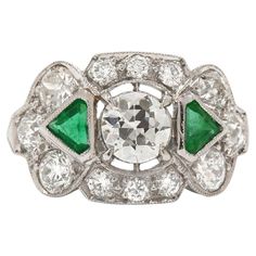 A unique Art Deco diamond and emerald three stone engagement ring. Centered by a captivatingly brilliant 1 carat old European diamond flanked by a pair of lush, vivid green emerald triangles. As if that wasn't enough, this 1920s Jazz Age heirloom is accented with a dozen dazzling round diamonds to complete this trinity band. Already have a wedding ring? Consider this as a stunning cocktail ring or a dynamic statement piece. Photography © Bella Rosa Galleries Santa Barbara, CA. Condition: Very Good Era: Art Deco Year: 1915-1935 Metal: Platinum Center Diamond: Carat Weight: 0.95 Cut: Old European Brilliant Color: I Clarity: VS1 Accent Diamonds (12): 3.23 carat total weight Color: G-H Clarity: VS1 Emeralds (2) Cut: Triangle Step Cut 0.50 carat total weight Ring Size: 7 1/4, sizeable Rise Abov Emerald 3 Stone Engagement Ring, Art Deco Engagement Ring Emerald, Three Stone Engagement Rings Emerald, Cluster Ring Set, Authentic Art, Emerald And Diamond Ring, 3 Stone Engagement Rings, Jewelry Appraisal, Jazz Age