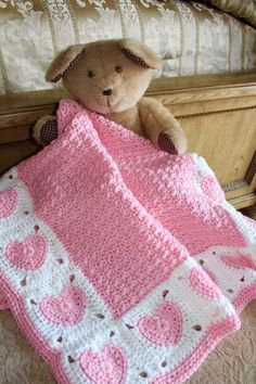 a teddy bear sitting on top of a bed next to a pink crocheted blanket