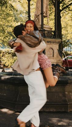 two people dancing in front of a fountain with trees and buildings behind them, one person has her arms around the other