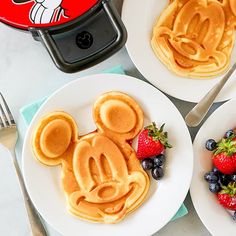 three mickey mouse waffles with strawberries and blueberries on plates next to a pancake