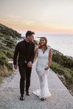 a man and woman walking down a path next to the ocean at sunset, holding hands