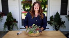 a woman standing in front of a wooden table holding a basket filled with flowers and plants