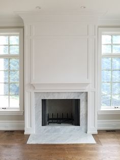 an empty living room with a fireplace and two windows in the corner, all white