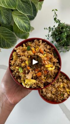 someone is holding two small red bowls filled with rice and veggies on the table