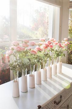 several white vases with pink flowers in them on a table next to a window