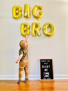 a little boy standing in front of some balloons that say big bro on the wall