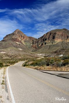 an empty road in the desert with mountains in the background and blue skies above it