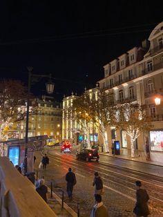 people walking down the street at night with buildings on either side and cars driving by