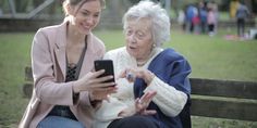 two women sitting on a park bench looking at a cell phone and pointing to the screen