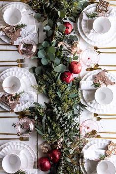 the table is set with white plates and silverware, greenery and red berries