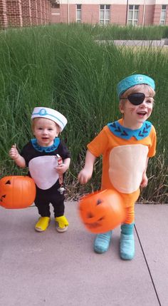 two young children dressed in costumes and holding pumpkins, standing on sidewalk next to tall grass