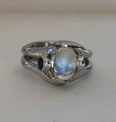 The Crystal Ball Ring is a very unique design, crafted by Heirloom Pavé in 18K white gold, featuring a Cabochon Moonstone. And two perfectly blue round brilliant Sapphires. The Moonstone is a oval shape Cabochon with a beautiful iridescent that so mystical and other worldly looking. The two, bezel-set Sapphires are a total carat weight of .35ct are a beautiful accent to the center. In the high polished white gold it is a true statement piece. Size 7 (Sizable upon request) Every Antique piece of Fusion Style Oval Moonstone Ring Gift, Oval Moonstone Fusion Ring Gift, Unique Opal Ring With Moonstone, Handmade Celestial Silver Opal Ring, Adjustable Silver Moonstone Multi-stone Ring, Unique Moonstone Wedding Ring, Mystical Cabochon Jewelry For Anniversary, Unique Silver Oval Cabochon Moonstone Ring, Unique Silver Moonstone Ring Oval Cabochon