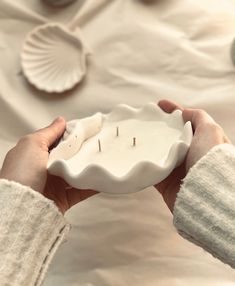 a person holding a small white bowl with candles in it and seashells on the table