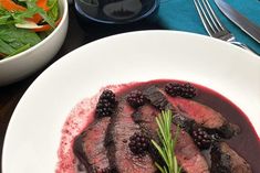 a white plate topped with meat and blackberries next to a bowl of salad on top of a wooden table