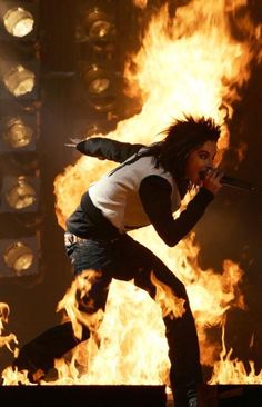 a woman in white shirt and black pants on stage with fire behind her at a concert