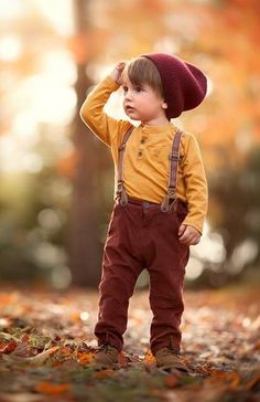 a little boy standing in the leaves wearing suspenders and a hat