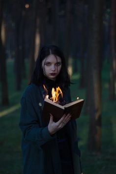 a woman standing in the woods holding an open book with flames coming out of it