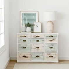 a white dresser with many drawers and pictures on the top, next to a lamp