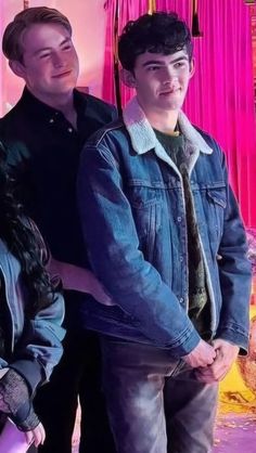 two young men standing next to each other in front of a pink wall with curtains