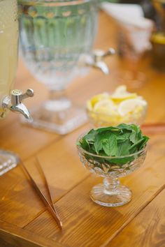 a wooden table topped with glass bowls filled with vegetables and lemon wedges next to a drink dispenser