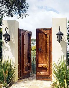 two wooden doors open in front of a white building with plants and trees on either side