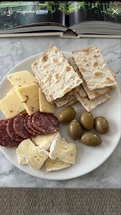 a white plate topped with crackers, olives and cheese next to an open book