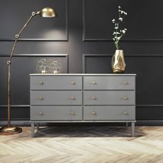 a grey dresser with gold accents and a lamp on top, against a black wall