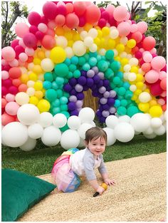 a little boy sitting on the ground in front of a rainbow balloon arch