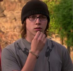 a man wearing glasses and a beanie talking on a cell phone while standing in front of a rock wall