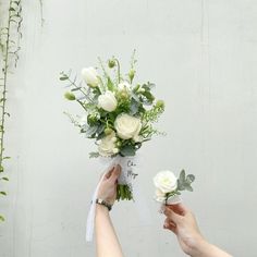 two hands holding white roses and greenery in front of a gray wall with vines