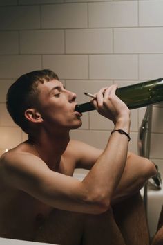 a shirtless man sitting in a bathtub drinking from a wine bottle